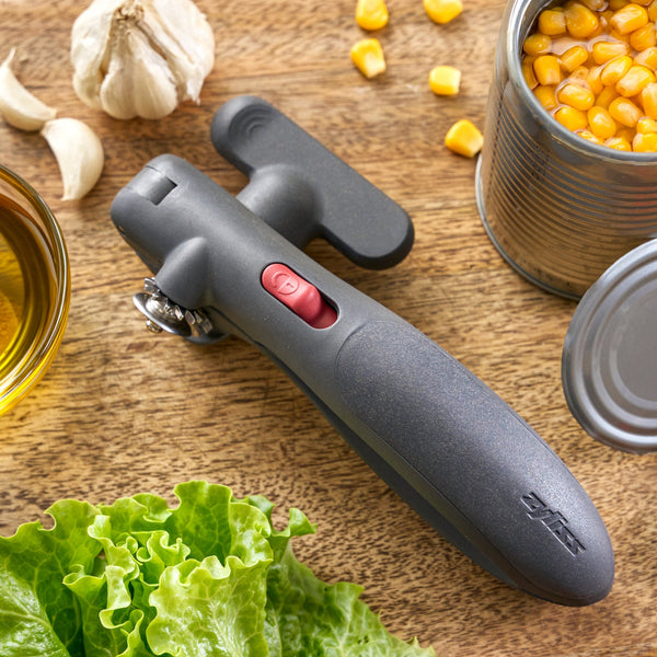 A manual can opener with a gray handle and red switch, shown on a wooden tabletop surrounded by food props including a can of corn, garlic, lettuce, and oil.