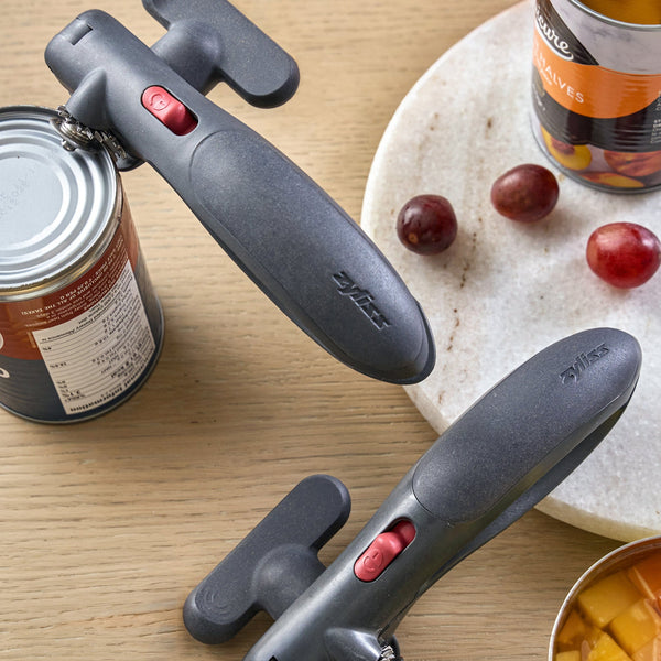 Two black manual can openers with red switches are shown on a wooden tabletop, with canned fruit and grapes used as contextual props.
