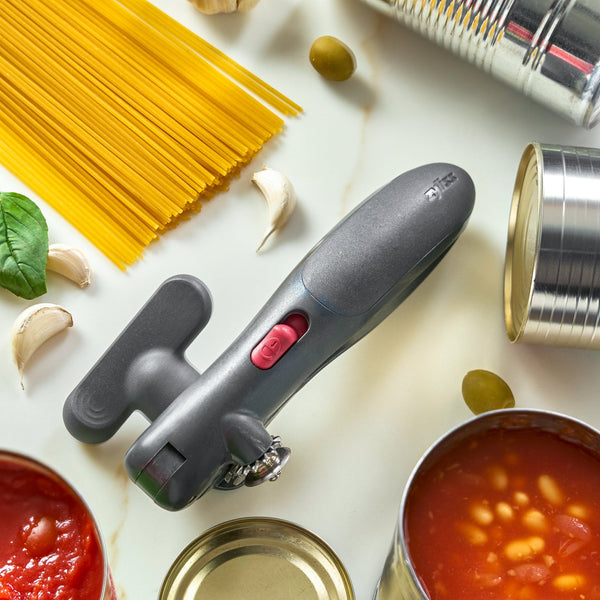 A gray manual can opener with a red switch is arranged on a white surface surrounded by cooking ingredients and canned goods, including spaghetti, garlic, olives, tomatoes, beans, and other open metal cans.