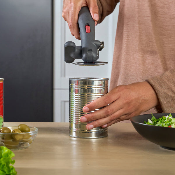 A person using a manual can opener to open a metal can on a kitchen counter, with canned olives, salad, and other kitchen items nearby.