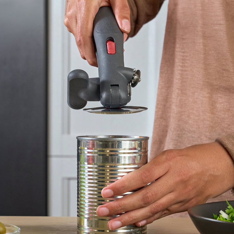 A person using a manual can opener to open a metal can on a kitchen counter, with canned olives, salad, and other kitchen items nearby.