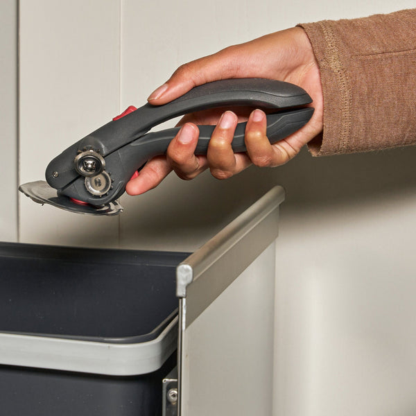 A hand holding a black and gray manual can opener above a dark gray trash bin.