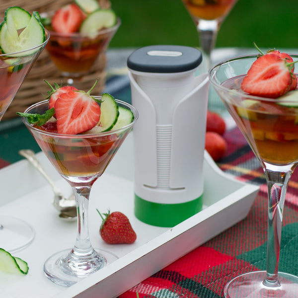 Four martini glasses with drinks garnished using spiral-cut strawberries, cucumber, and mint created with the Zyliss Spiralizer sit on a white tray atop a red plaid picnic blanket near a white insulated flask. 