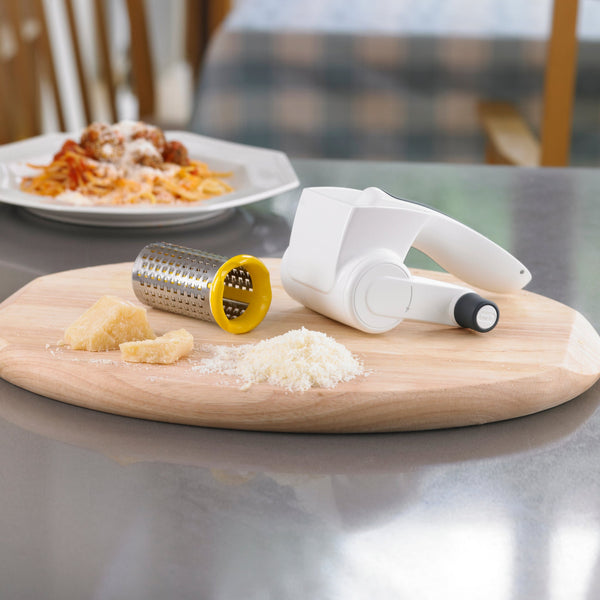 The Zyliss Classic Cheese Grater, featuring a stainless steel drum and ergonomic handle, sits on a wooden cutting board with grated cheese and cheese chunks nearby, while a plate of pasta rests on the dining table in the background. 