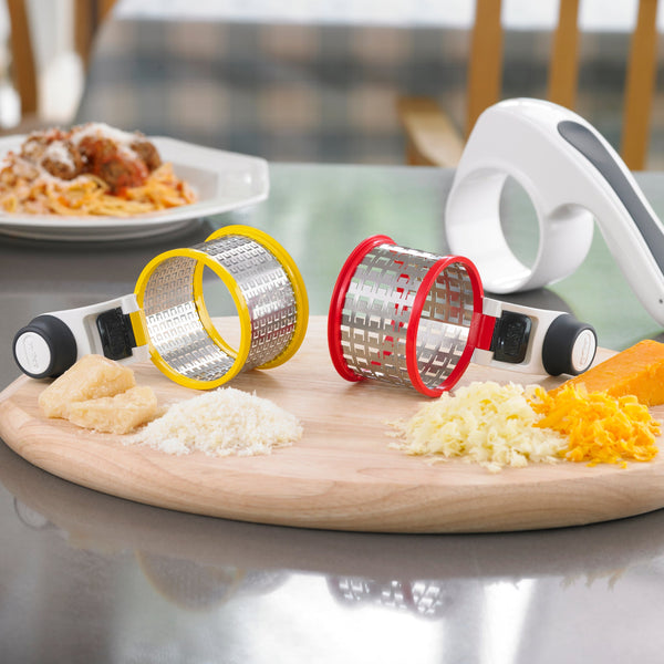 The Zyliss All Cheese Grater, featuring interchangeable drums and stainless steel blades, rests on a wooden board amid grated cheese and other foods, with a plate of pasta in the background. 