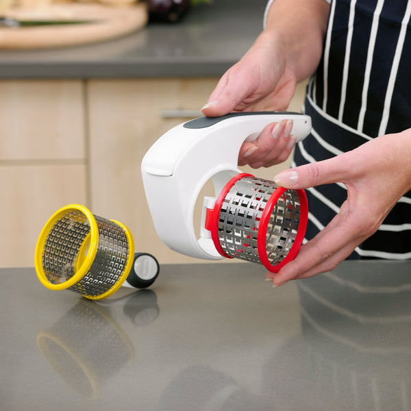 A person in an apron holds the All Cheese Grater by Zyliss, featuring sharp stainless steel blades and a white body with a red interchangeable drum, while a yellow drum attachment sits on the kitchen counter. 