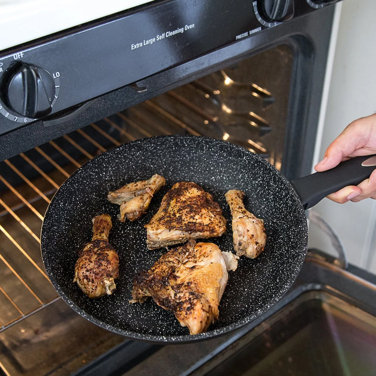 A hand holds a frying pan with cooked, seasoned chicken pieces in front of an open oven. The oven door is partially open, and the chicken appears to be ready for baking or finishing in the oven.