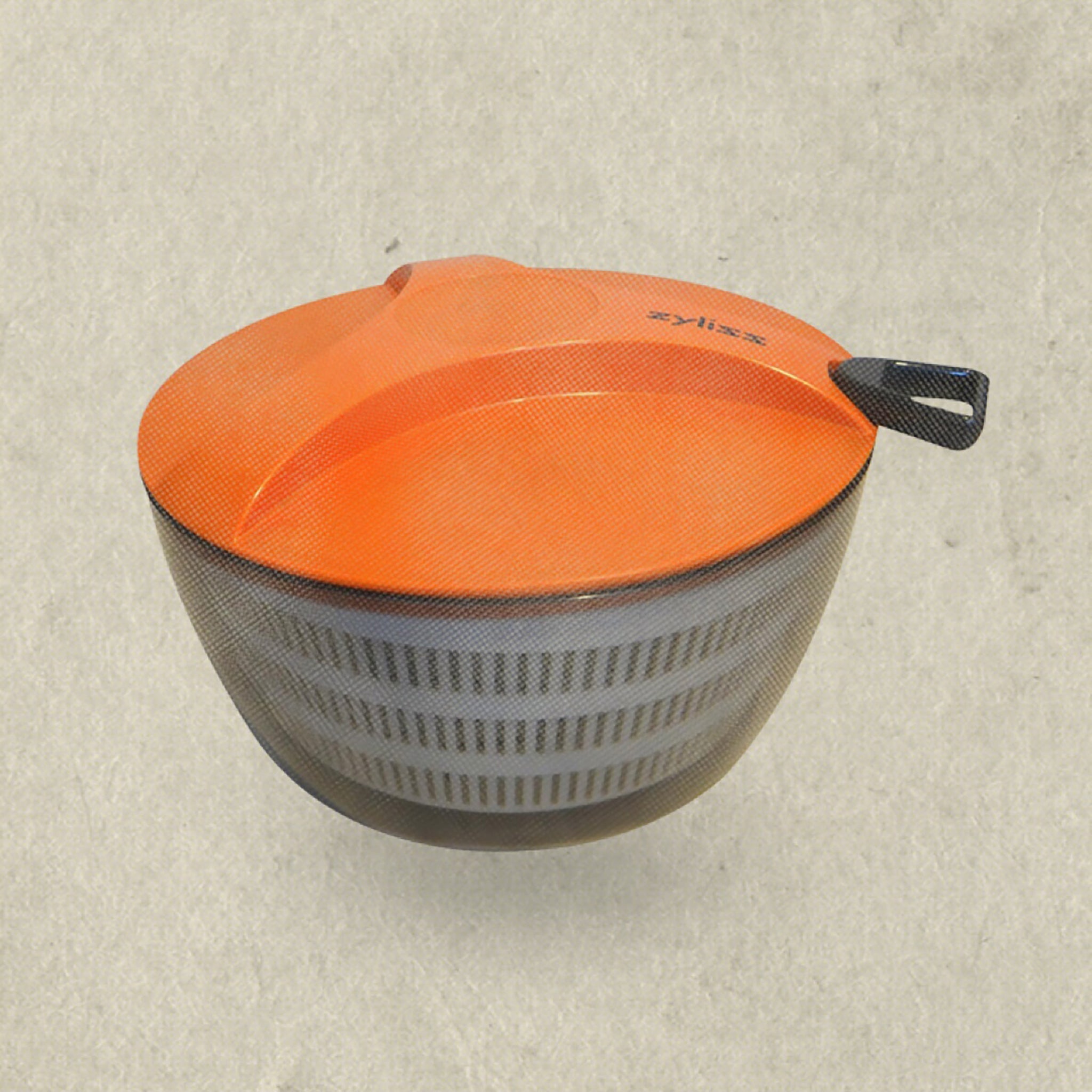 A salad spinner with a translucent bowl and a bright orange lid, featuring a black handle on one side, sits on a neutral textured background.
