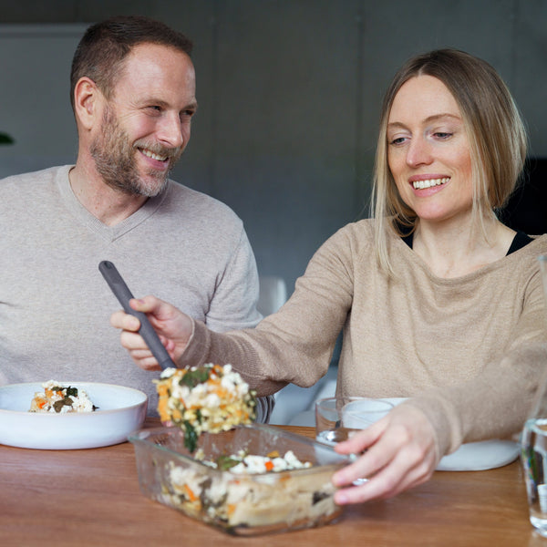 A woman serves a casserole from a Zyliss Glass Container while a man smiles at her. They enjoy meal prep together at a wooden table, with additional Zyliss glass storage containers nearby in a cozy, casual setting. #1.05L/1.1qt