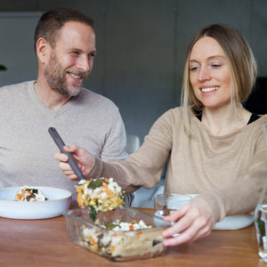 A woman serves a casserole from a Zyliss Glass Container while a man looks on. They sit at a wooden table, sharing meal prep in a cozy indoor setting, highlighting the secure and stylish Zyliss Glass Container. #1.52L/1.61qt