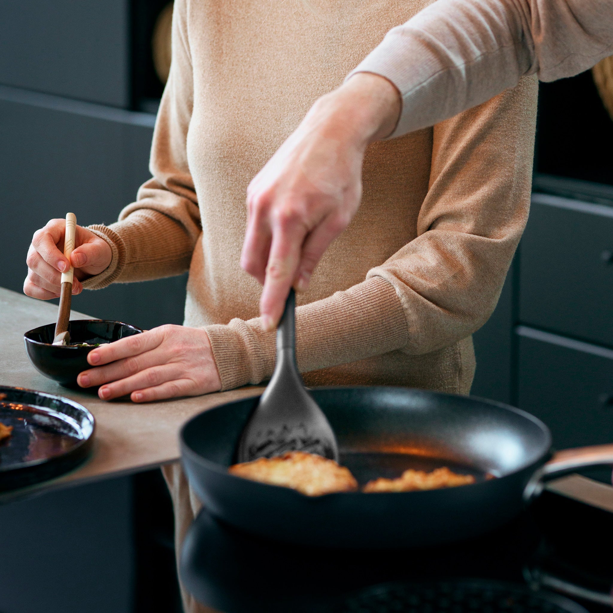Two people cook together; one stirs a small bowl, while the other uses the Zyliss Slotted Turner with an ergonomic handle to cook food in a frying pan on the stove. Their faces arent visible.
