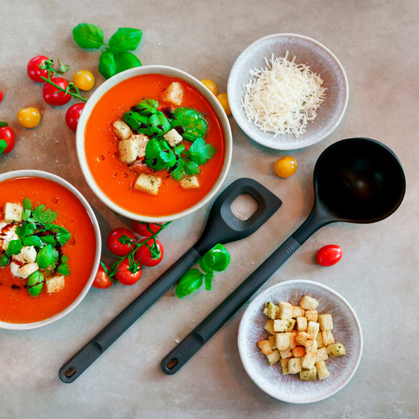 Two bowls of tomato soup topped with croutons and herbs, surrounded by cherry tomatoes, shredded cheese, a bowl of croutons, fresh basil, and two Zyliss Angled Mixing Spoons with ergonomic handles on a light surface. #Large