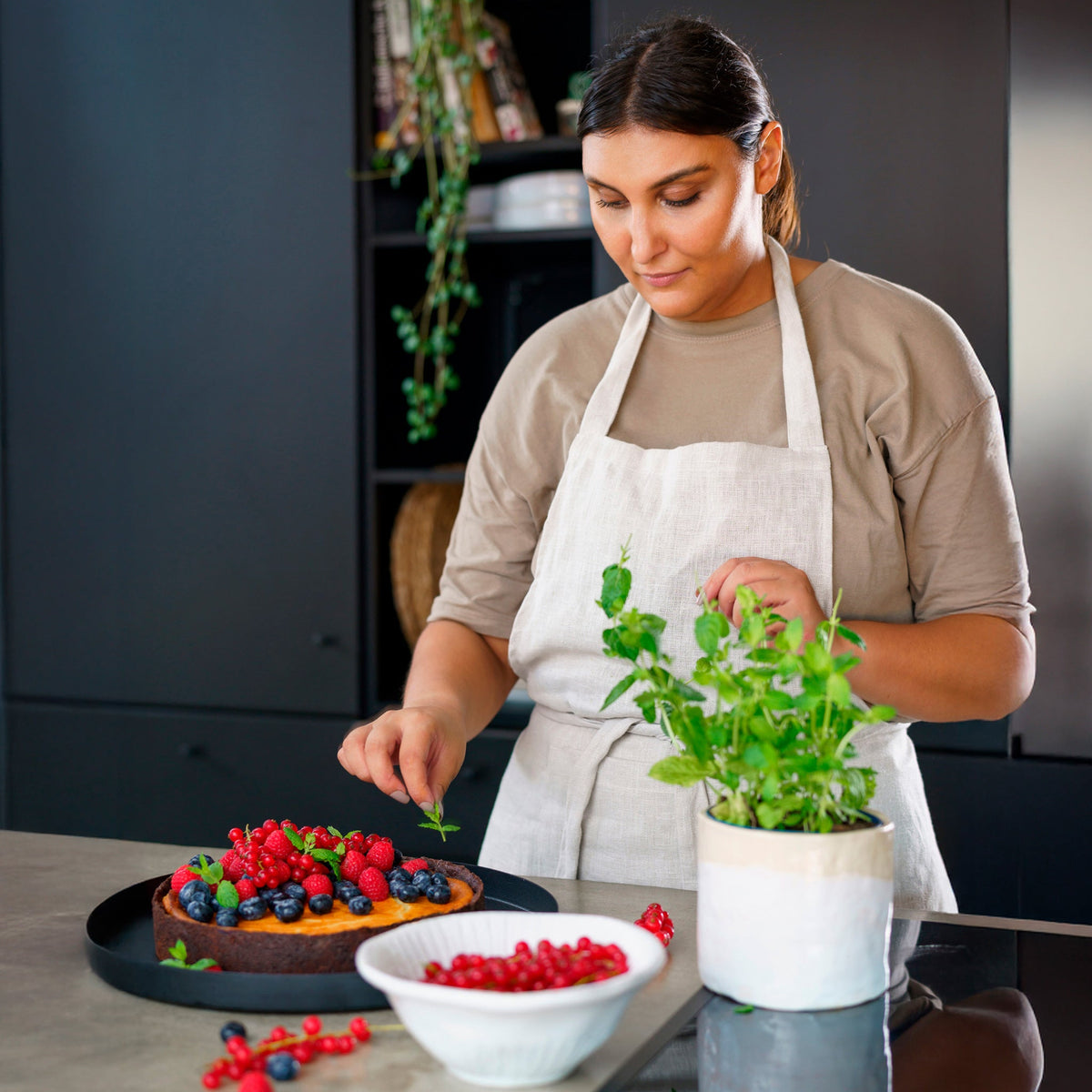 In a modern kitchen, a woman in a beige apron decorates a berry-topped cake with mint using the durable Zyliss Spoon. A bowl of red currants and a potted herb plant rest on the counter nearby. #Large